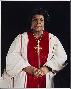 Deborah Wolfe posing for a portrait in red and white robs, wearing a golden cross, hands crossed in her lap.