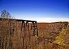 A trestle bridge across an autumnal valley at left ends in a drop off at center, with collapsed remnants at right, all under a bright blue sky