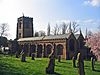 A substantial red sandstone church seen from the southeast. The tower and body of the church are crenellated. In the churchyard are gravestones and at the right extremity is a tree in flower