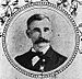 Head and shoulders of a white man with a wide bushy mustache, wearing a bow tie and dark suit with a medal pinned to the left breast. The portrait is surrounded by a circular frame and images of flowers.