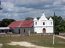 An open field with patchy, worn grass extending towards a low church with a simple white facade. The facade has three short spires, with the middle spire taller than the flanking spires. The central spire supports a cross. The shorter spires sit above arched openings, a bell is visible in the left-hand arch. An arched wooden door is set into the facade; a square window is above it. The main body of the church only stands about half the height of the facade and extends back to the left. The wall is bare except for a smaller doorway and a small window. The roof is rusty corrugated metal. A lower building with a shaded balcony extends to the left of the church. Two poles support a power line running across the field to the church. The base of each pole is painted white. A line of trees runs across behind the church.