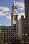 Distant, ground-level view of white limestone tower. The building is topped with a pyramidal-cap, and a large, 4-faced clock is visible just below the main roofline