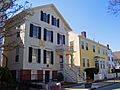 Two houses stand near the street, with no front yard. The left house is a two story white colonial, and the right one is also a two story colonial, painted yellow.