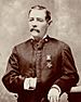 A white man with a mustache standing with his left arm resting on an object to his side and his right hand inside his jacket. A star-shaped medal is hanging from a ribbon on his left breast.