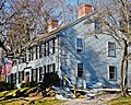 A blue colonial two story house with black shutters. Over the front door is a flag pole holding an American flag.