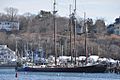 Adventure is a long black ship with three masts. She sits relatively low to the water, anchored at a berth in front of other smaller ships, and land rising in the background.