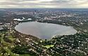 An aerial view of Bde Maka Ska, a mostly round lake with two nubby "legs" at its bottom end, surrounded mostly by parks and houses. Downtown Minneapolis is visible at the top of the image.