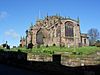 A red sandstone Perpendicular-style church seen from the east at a low angle. In the foreground is a wall, partly in shadow, behind which are daffodils and a vertical public footpath signpost