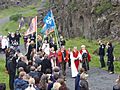 A crowd of people walking along an outdoor path. They are led by individuals in robes, and a number carry flag banners.