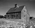 This black and white photo shows a saltbox-style house with split-rail fencing in front. A meadow with flowers is in the foreground, and small buildings associated with the house are in the background.