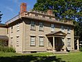 A large brown colonial frame house. There is a railing going around the roof, and a bumped up central section is visible in the center of the house. A portico shelters the front door.