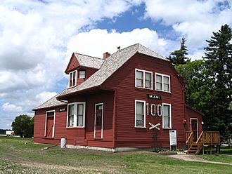 A small dirt path leads from the bottom right to the side of a red building and a five-step deck stairway at the end of which is a door. The side of the building is two stories, with two narrow windows on the bottom floor flanking the number 100, below which is a railroad crossing as a decoration and above which is a sign reading "Miami". Above these are two square windows. The building extends away to the left, with the steep roof pierced by a gable, beyond which is the roof of the single-storey freight shed. The walls consist of horizontal planks painted red, and the window frames are painted white. Behind the building are a few trees, with a cloudy sky in the background, and fronting the structure is green grass, patchy in some areas.