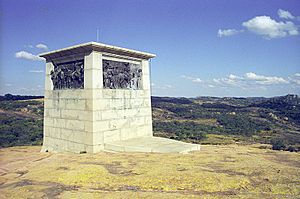 A white, oblong, flat-topped structure, with soldiers depicted on each side in bas relief