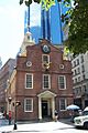 Ground-level view of a 3-story, colonial-era brick building with a prominent white tower on one end; several glass skyscrapers are visible in the distance.