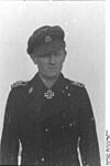 A man wearing a military uniform, peaked cap and a neck order in the shape of a cross. His cap has an emblem in shape of a human skull and crossed bones.