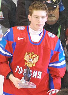 A short- and light brown-haired caucasian male in a red, Russian, ice hockey uniform is holding a small glass award.