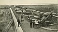 an elevated photo of five guns of three kinds in a row, with several people posed along a fort parapet