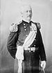 A white man with mustache, wearing an ornate military uniform including a sash across the chest, fringed shoulder boards, white gloves, and several medals pinned to the left breast.