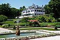 A white mansion house stands in a garden. A terraced lawn separates the house from a fountain with a pool fringed by white plants.