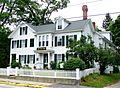 A rambling colonial house, painted white with black shutters. The front yard is surrounded by picket fencing, and a sign mounted on the front door's portico reads "Whittier Home".