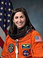 An astronaut wearing her orange mission suit with the American flag embroidered on her shoulder and name tag and mission patches embroidered on her chest.