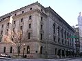 A stone building five stories high with a parapeted decorated flat roof seen from across an intersection, with traffic lights in the foreground. The windows along the right side are arched. There are taller buildings in the right background.