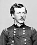 Head and shoulders of a young white man with wavy hair and a mustache, wearing a double-breasted military jacket with a rectangular patch over each shoulder.