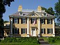 A large yellow house with white trim and black shutters. The front door is flanked by pillar-like decorative molding, and is sheltered by a small porch. The roof is ringed by a low railing.