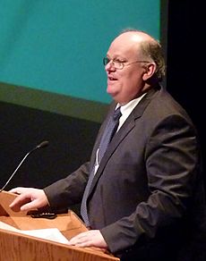 Man in his 50s wearing glasses and a suit standing at a lectern