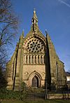 The west end of a Gothic Revival church with a small doorway, arcading, a large and elaborate rose window, and a narrow bellcote