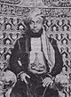 A black-and-white photograph of a man with a dark beard wearing a turban, a dark jacket, and a white shirt, sitting, and looking at the viewer