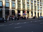 A large crowd of people walking on a grey sidewalk next to a black road where two vehicles are driving from the left to the right