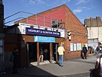 A red-bricked building with a rectangular, blue sign reading "HIGHBURY & ISLINGTON STATION" in white letters all under a blue sky with white clouds