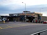 A building with a blue sign reading "BLACKHORSE ROAD STATION" in white letters and several people and cars in the foreground all under a blue sky