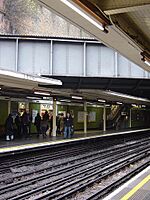 Eight people walking towards an escalator on a railway platform next to a green-tiled wall on the left and a railway track on the right