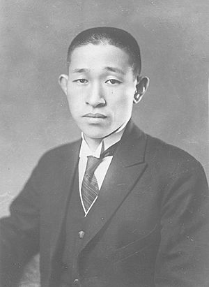 Black and white portrait. Head shot of Matsushita in front of a dark background, wearing a dark suit jacket with a light wing-collared shirt, dark vest, and dark striped necktie.