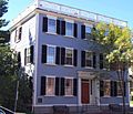 A three-story colonial-style house with a low railing around the roof. The house is gray with white trim and black shutters. There is a small portico sheltering the front door.