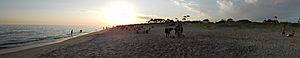 A panoramic photograph of a beach on a sunny day. There are people playing in the water, sitting in chairs, and walking in the sand along the water.