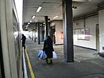 Three people walking on a platform, the one on the left boarding a train, the one in the middle carrying bags, and the one on the right walking through a door