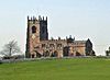 A sandstone church seen from the south at a distance standing on a mound. The tower to the left has crocketted pinnacles and the nave and aisle are crenellated. The foreground consists of a grassy field