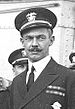 Head and shoulders of a white man with a handlebar mustache, wearing a peaked cap and a dark jacket with two rows of ribbon bars on the left breast. Several other men in similar clothing can be seen behind him.