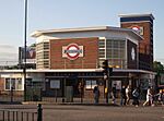 A red-bricked building with a blue sign reading "BOUNDS GREEN" in white letters and several people walking in the foreground all under a blue sky