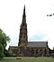 A sandstone church with a very tall spire and a protruding transept seen from the south. In front of the church is a grassed area containing a bench and to the left is a tree