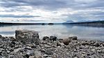 A wide flat river flows along a rocky shore toward a mountain range in the far distance.