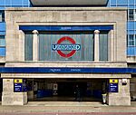 A grey building with a rectangular, dark blue sign reading "MORDEN STATION" in white letters, two red buses in front, and a fence in the foreground