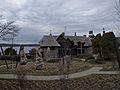 A stone and wooden house with a wooden roof stands behind a garden with plants wrapped in burlap for the winter. The land slopes down to water behind the house.