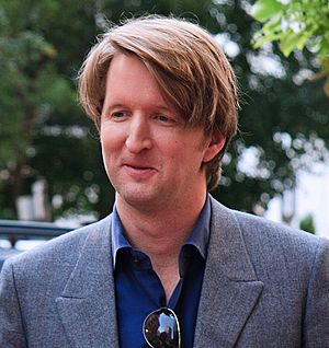 A man in a grey suit with a blue shirt looks down to his right while smiling.