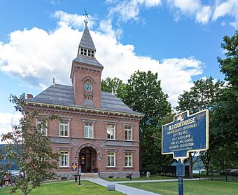 Front view of old courthouse building, with a brick clock tower, pointed slate roof, and plaque in front