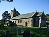 A small church seen from the southeast. The tower has a small pyramidal spire, a porch protrudes from the south wall, and the chancel to the right is short and slightly lower than the nave. Gravestones in shadow are in the foreground