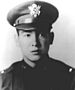 Head of a young man wearing a peaked cap with a large, shiny emblem on the front and a dark military jacket.